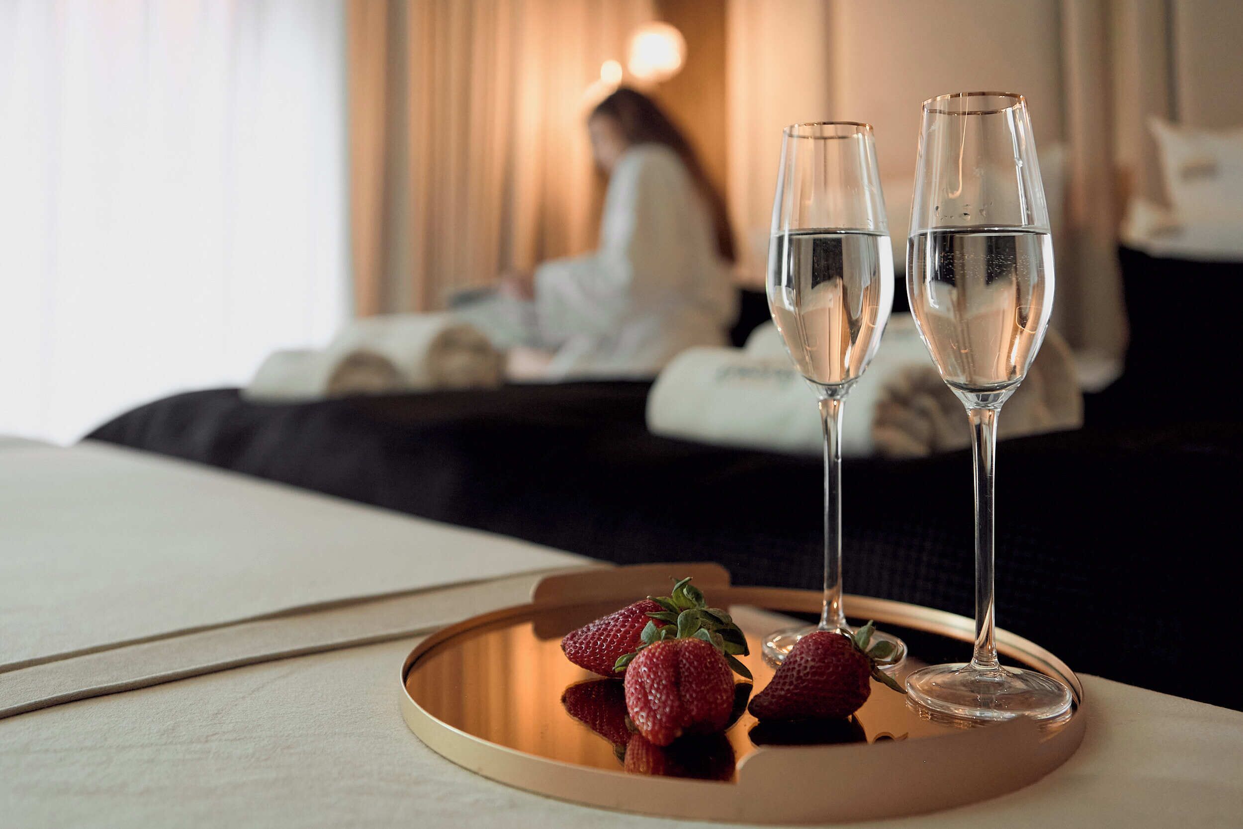 A hotel room with a bed, a bedside table with two champagne flutes and strawberries, and a person in the background.