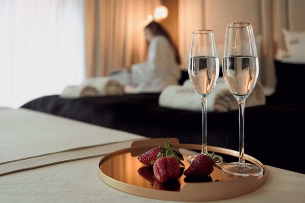 A hotel room with a bed, a bedside table with two champagne flutes and strawberries, and a person in the background.