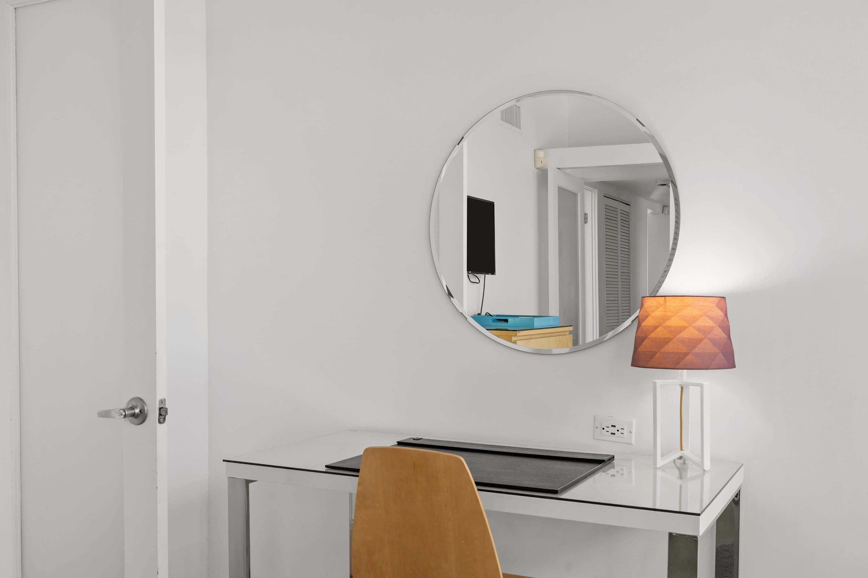 Una habitación con una puerta blanca, un espejo redondo, un escritorio con una computadora portátil y una lámpara.