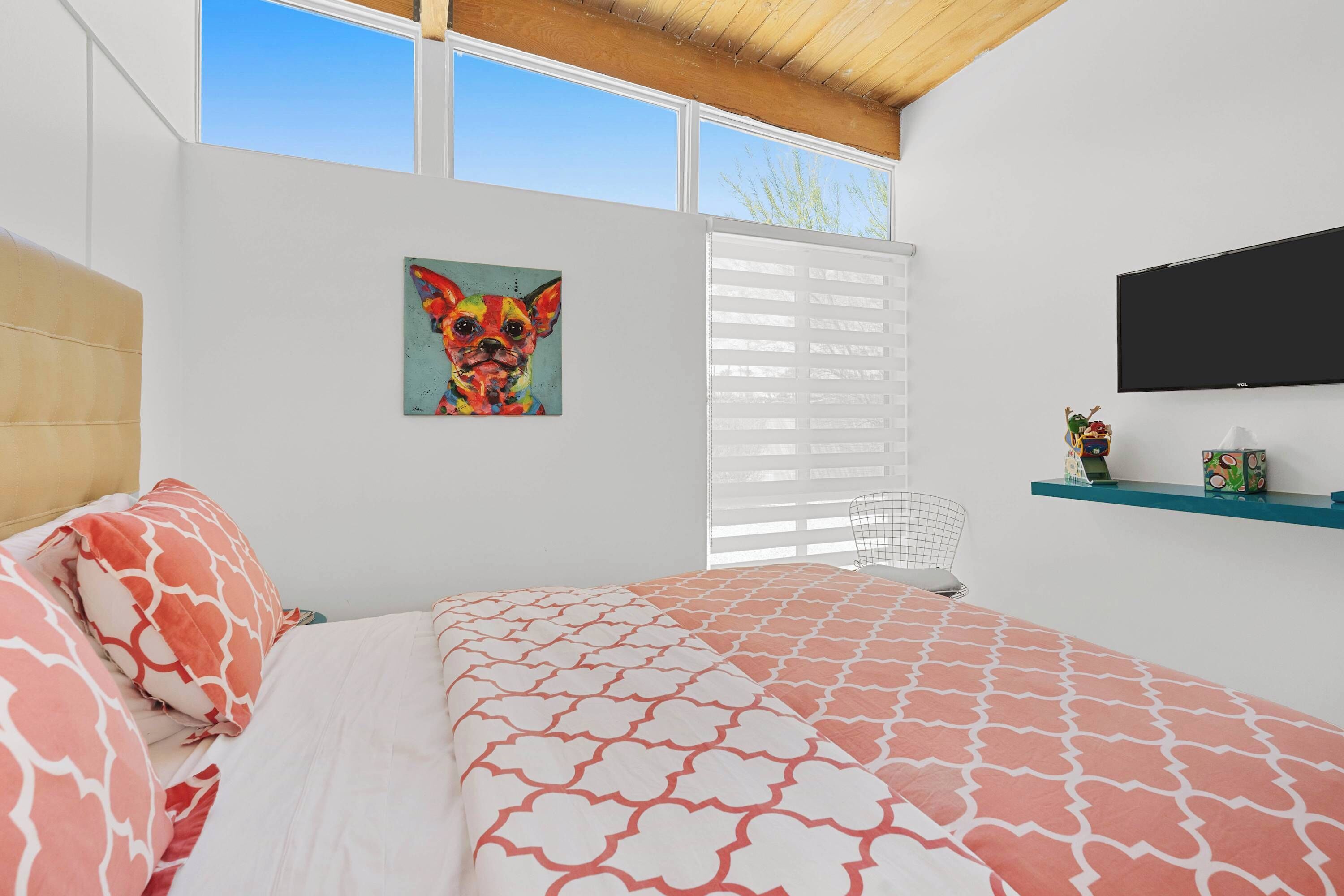 Une chambre à coucher avec un lit, un tableau représentant un chien, une télévision et une fenêtre avec des stores.