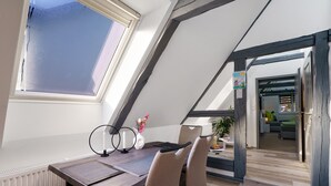 A modern loft bedroom with a sloped ceiling, a skylight, a dining area with a table and chairs, and a view into the living room.