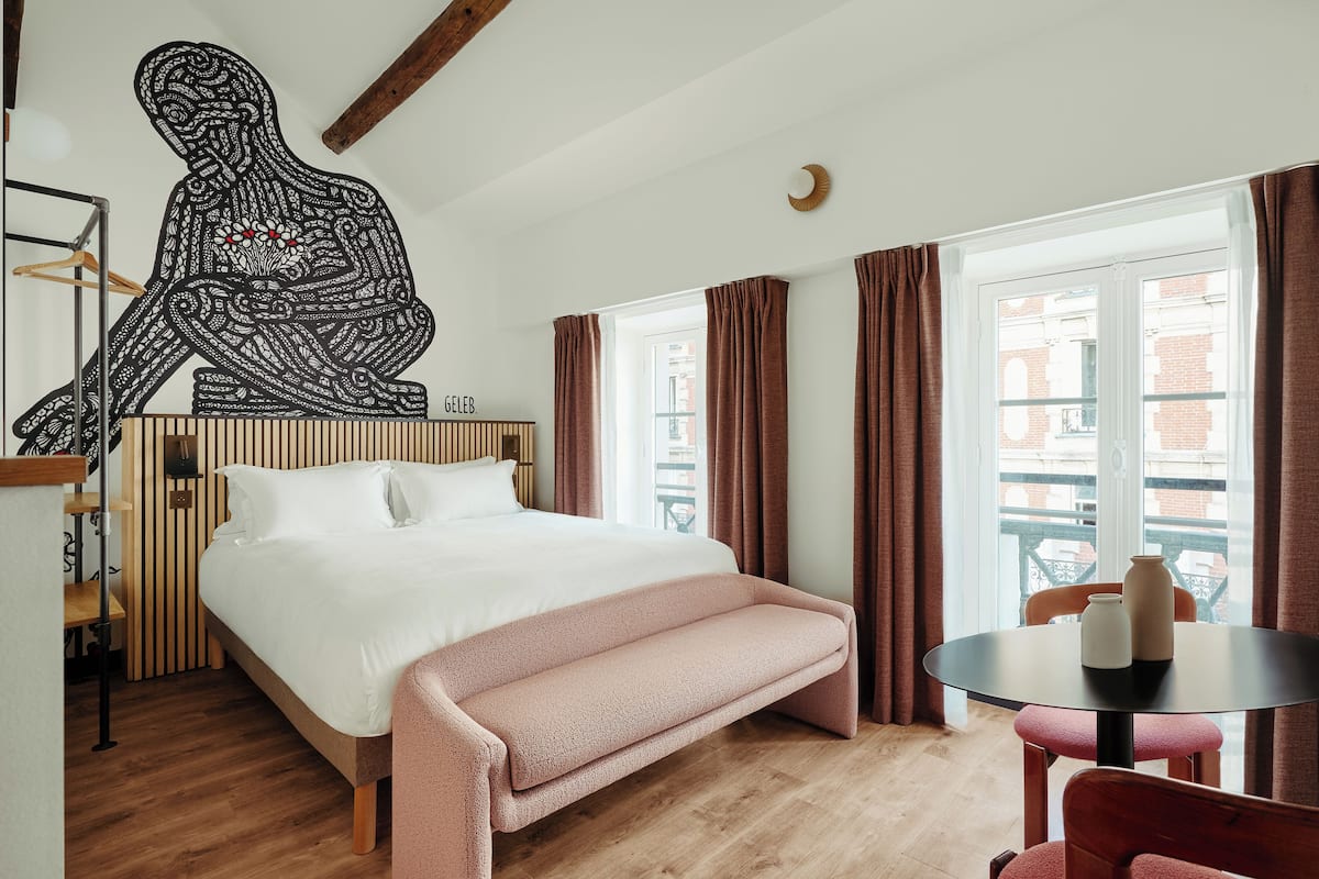 Quarto com cama, um banquinho, uma mesa com vasos e uma obra de arte na parede.