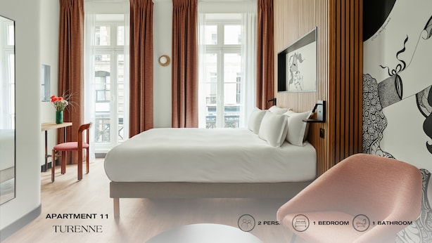A modern bedroom with a large bed, a desk, and a chair. There are two windows with curtains, a wall-mounted TV, and a decorative wall piece. The room has a wooden ceiling and a view of buildings outside. - Beauquartier - Marais Turenne (Paris)