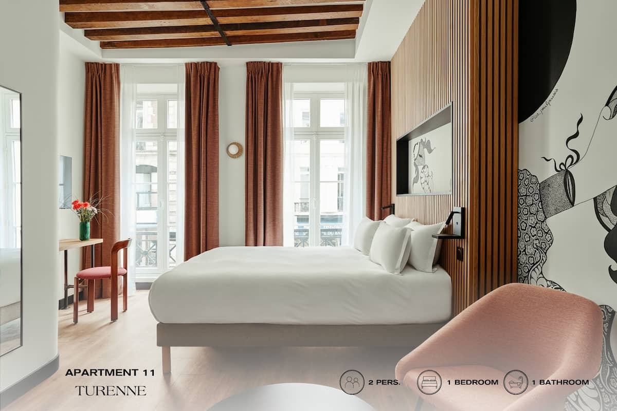 Quarto moderno com cama grande, escrivaninha e cadeira. Há duas janelas com cortinas, uma TV fixada na parede e um objeto decorativo. O quarto possui teto de madeira e vista para edifícios.