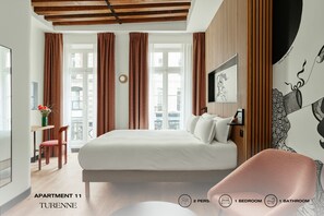 A modern bedroom with a large bed, a desk, and a chair. There are two windows with curtains, a wall-mounted TV, and a decorative wall piece. The room has a wooden ceiling and a view of buildings outside. - Beauquartier - Marais Turenne (Paris)