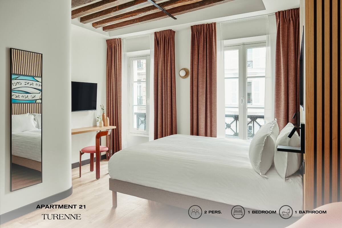 Quarto moderno com cama, escrivaninha e TV. Cortinas, janela e vista para o exterior. As paredes são brancas e o teto é de madeira.
