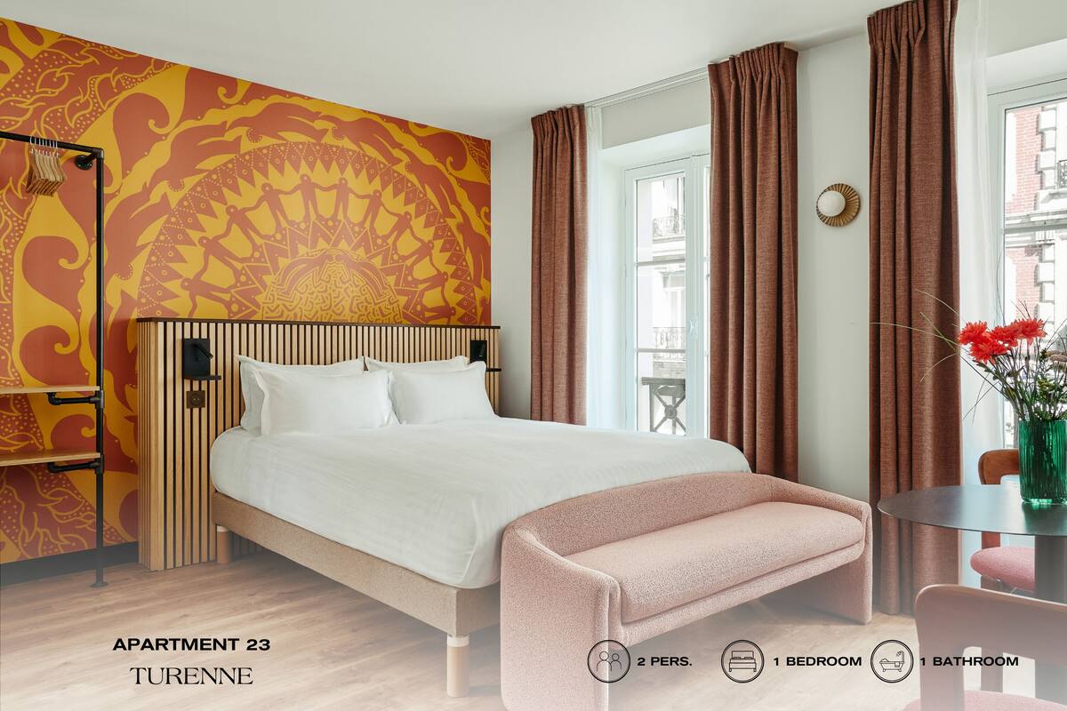 Quarto com cama, um banco, uma mesa com flores, uma janela e parede estampada. Identificado como Apartamento 23, Turenne, para 2 pessoas, 1 quarto e 1 banheiro.