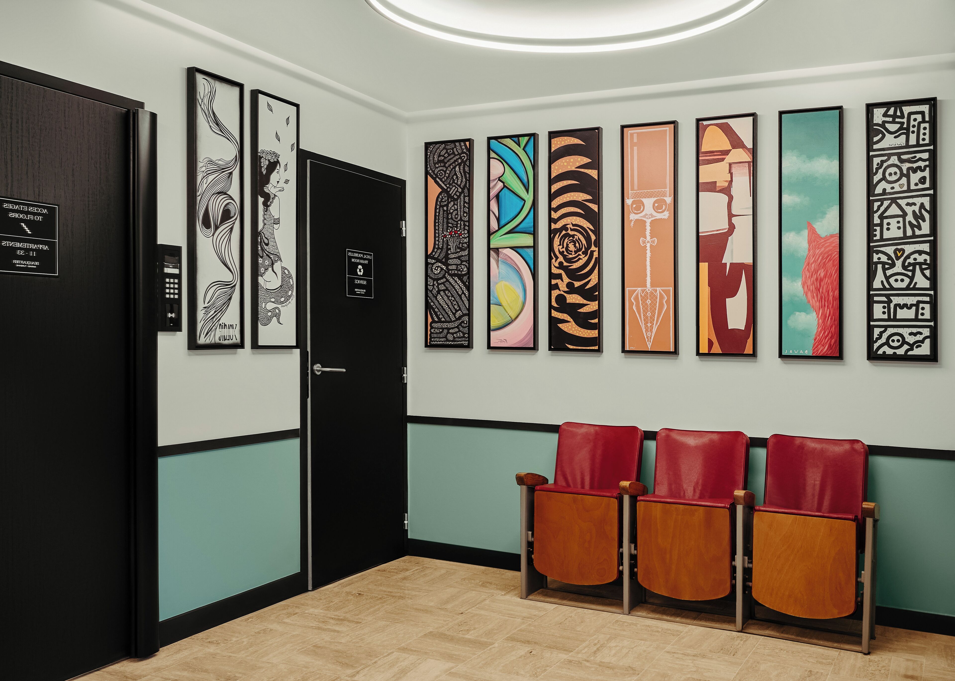 A hallway with a black door, a row of framed artworks on the wall, and a seating area with red chairs.