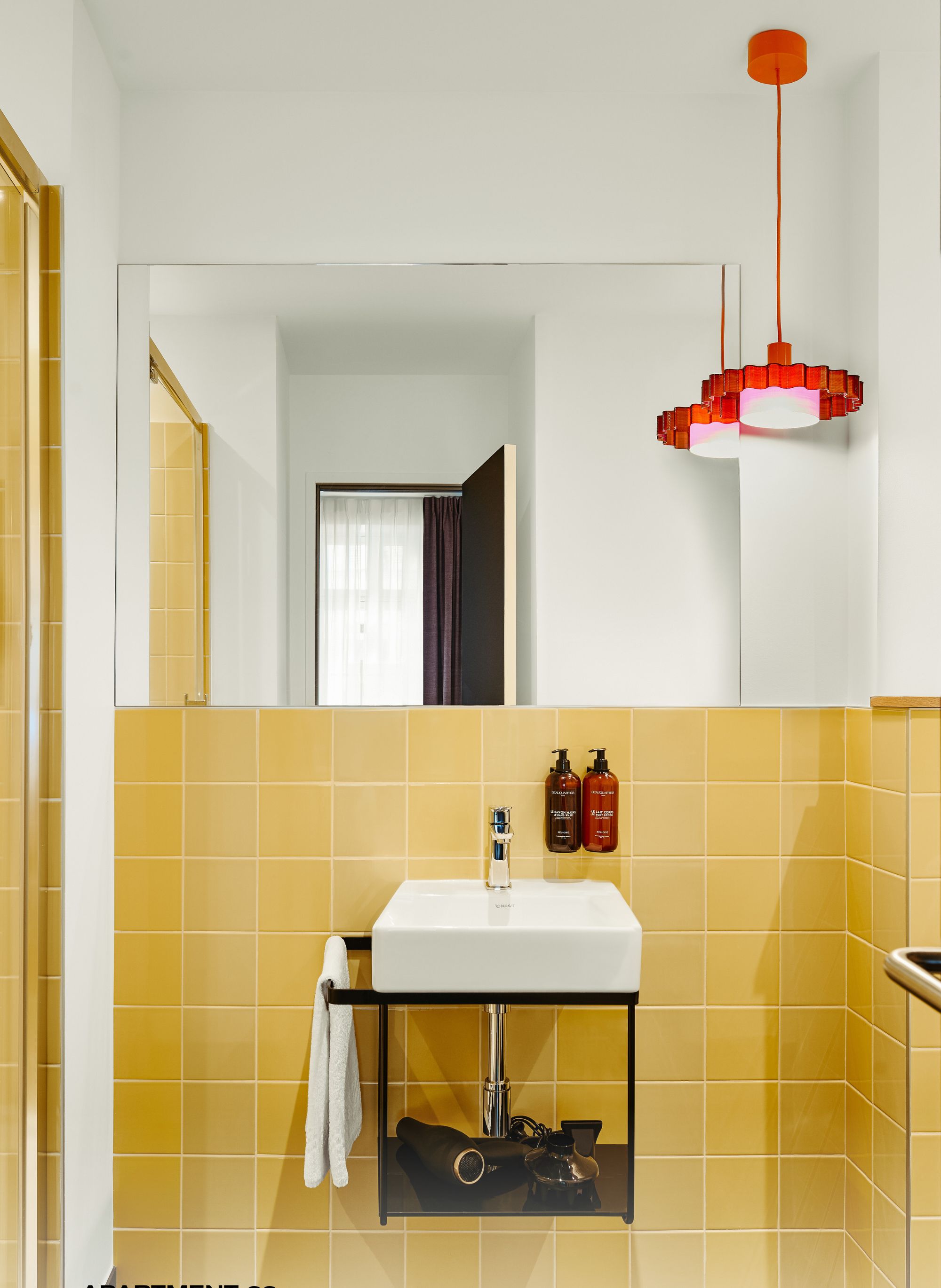 Een badkamer met gele tegels, een witte wastafel en een rode hanglamp. Er staan twee flessen op de muur en een handdoek hangt aan de rek.