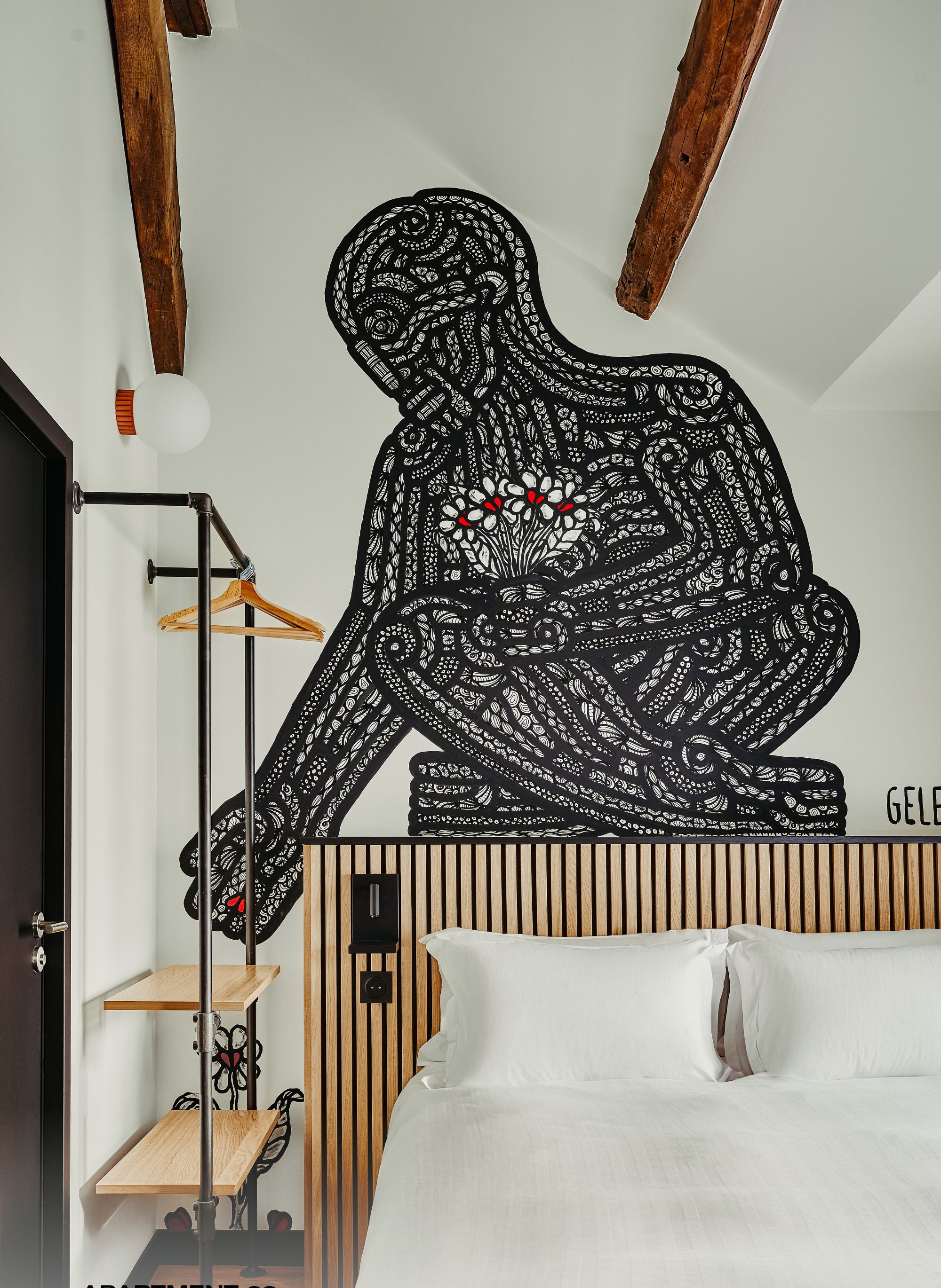 Ein Schlafzimmer mit einem Bett, einem hölzernen Kopfteil, einer Pendelleuchte, einem Kleiderständer und einem Wandbild.