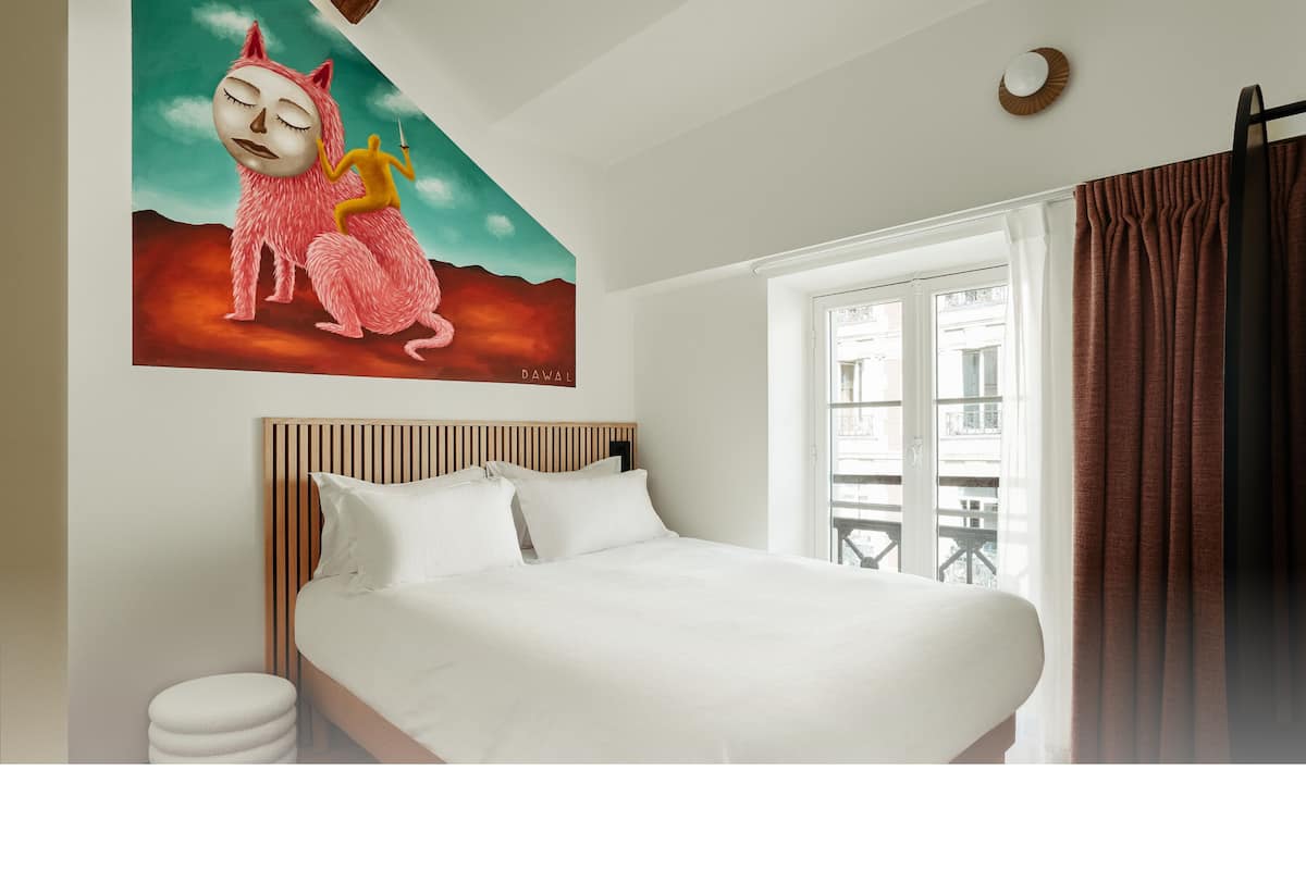 Quarto de hotel com uma cama grande, cabeceira de madeira, travesseiro branco, mesa de cabeceira redonda e uma janela com cortinas.