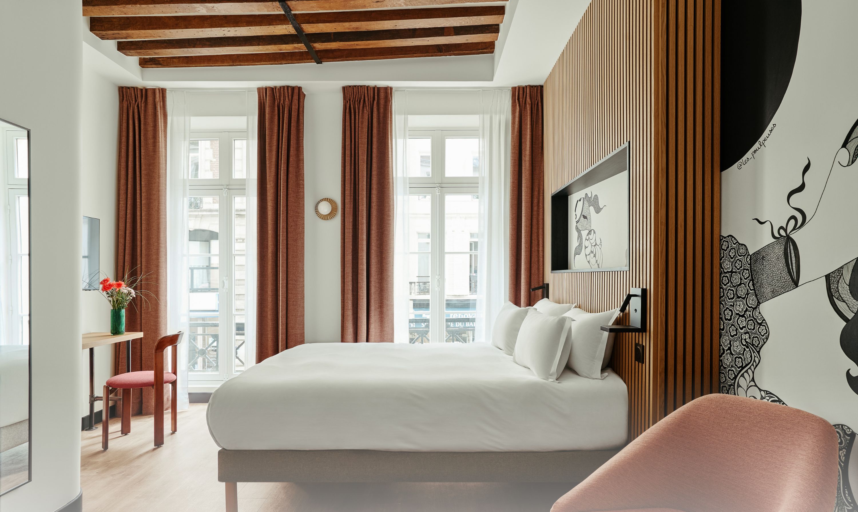 A modern bedroom with a large bed, a desk, and a chair. There are two windows with curtains, a wall-mounted TV, and a decorative wall piece. The room has a wooden ceiling and a view of buildings outside.