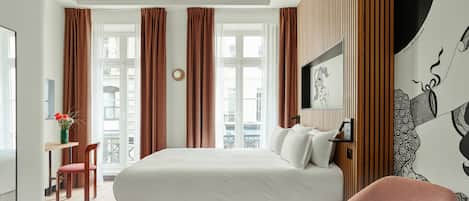 Une chambre moderne avec un grand lit, un bureau et une chaise. Elle est dotée de deux fenêtres avec des rideaux, d’un téléviseur fixé au mur et d’un élément décoratif mural. La chambre a un plafond en bois et offre une vue sur les bâtiments environnants.