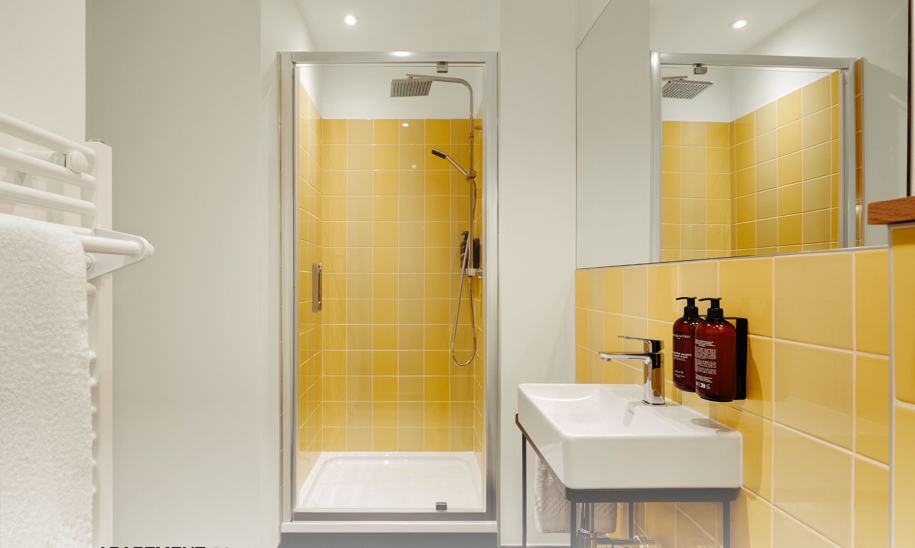 Un baño moderno con azulejos amarillos, ducha envuelta en vidrio, lavamanos blanco y espejo.
