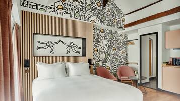 Un dormitorio moderno con cama, una silla, una mesita pequeña y una pared con un diseño único. Hay una puerta que da a un baño y una kitchenette. La habitación tiene un techo de madera y un panel de pared a rayas.