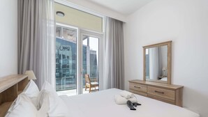 Hotelkamer met een groot bed, een houten dressoir, een spiegel en uitzicht op een stadsgezicht door raamhoog plafond ramen.