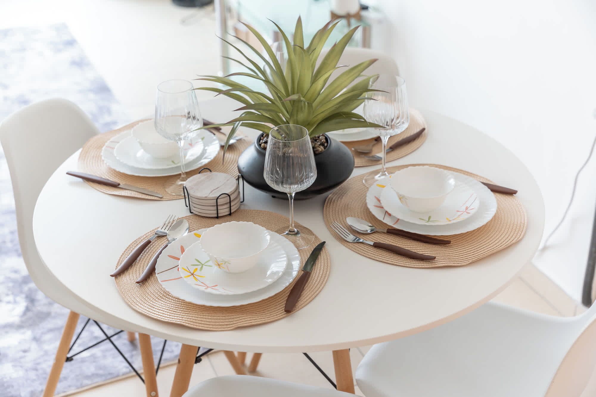 Een ronde eettafel gedekt met borden, bestek en glazen, met een potplant in het midden.