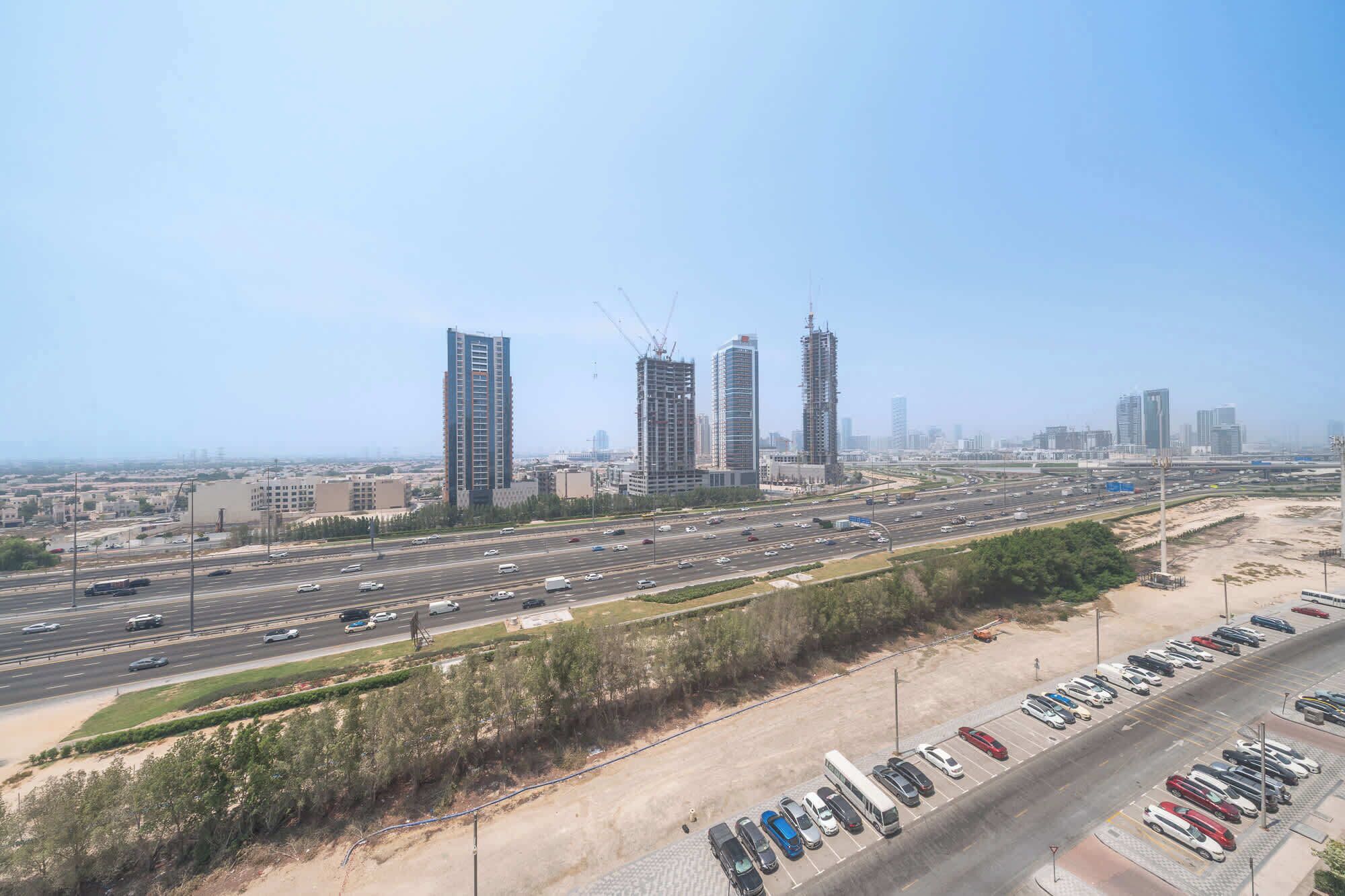 A cityscape with multiple high-rise buildings, including some under construction, and a busy highway with a parking area in the foreground.