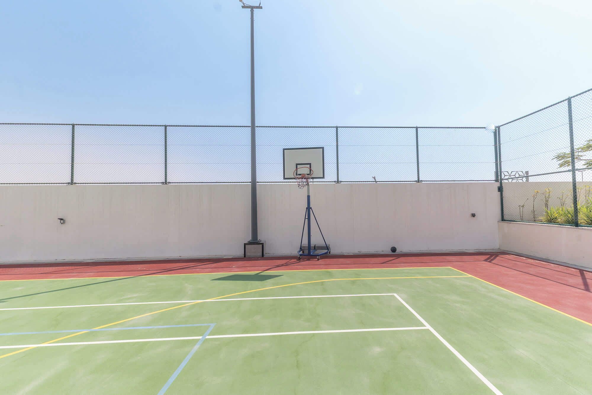 Una cancha de baloncesto al aire libre con un aro, rodeada por una cerca de malla metálica y un cielo despejado.