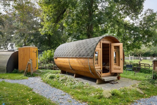 A wooden barrel-shaped cabin with an open door, a wooden ladder, and a gravel path.