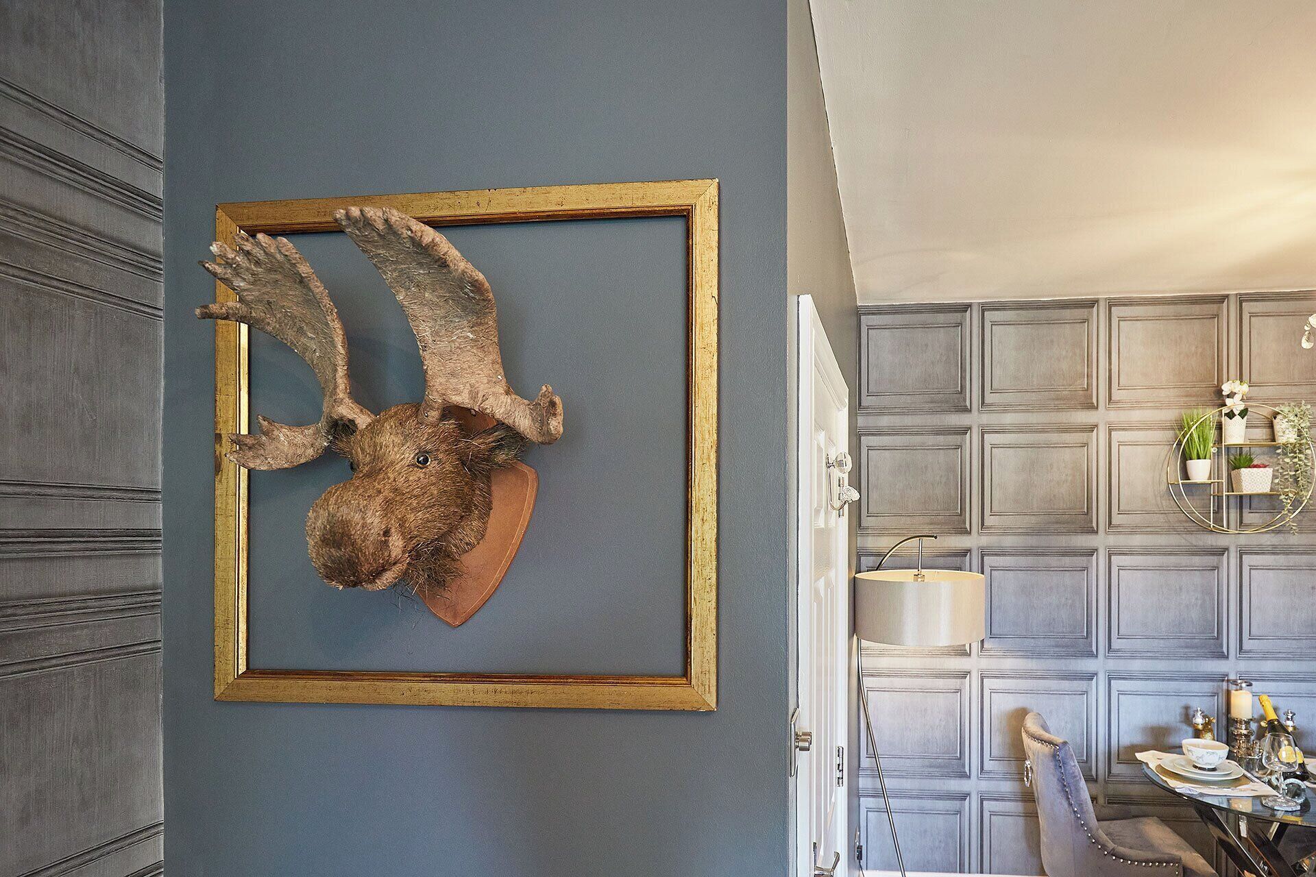 A room with a mounted animal head in a gold frame on the wall, a dining area with a table and chairs, and a decorative wall paneling.