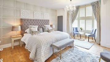 A bedroom with a large bed, a bench, two chairs, a small table, and a chandelier.
