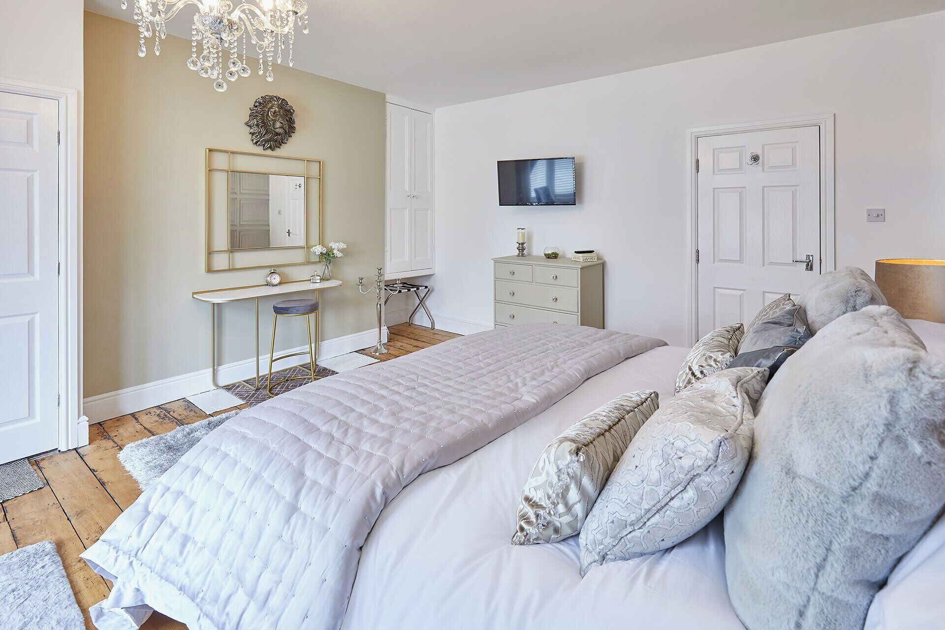 A bedroom with a large bed, a vanity with a mirror, a television on the wall, and a chandelier.