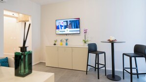 A modern hotel room with a flat-screen TV mounted on the wall, a small round table with two black bar stools, a beige cabinet, and a potted plant. - Imperium Suite Trevi (Rome)