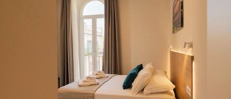A hotel room with a bed, a bedside table, a window with curtains, and a wall-mounted light fixture.