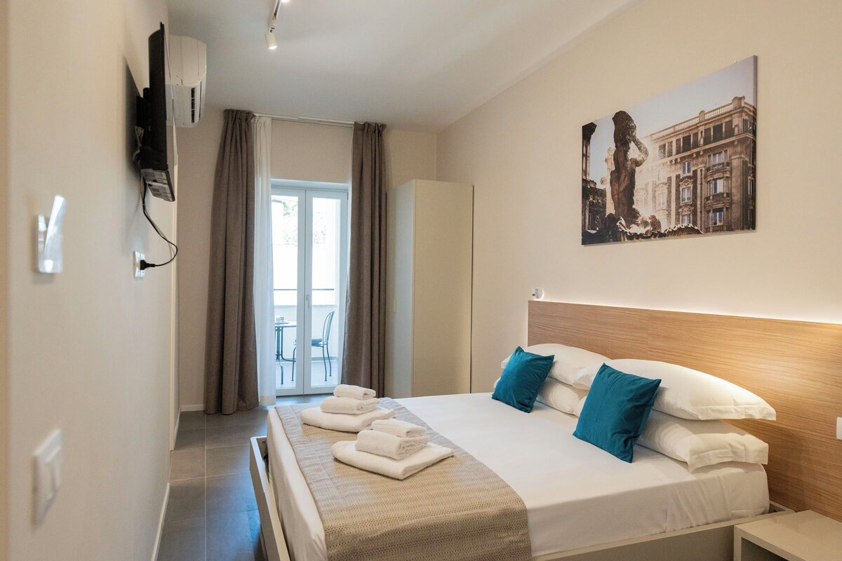 A hotel room with a bed, a painting, a TV, and a view of the outside.