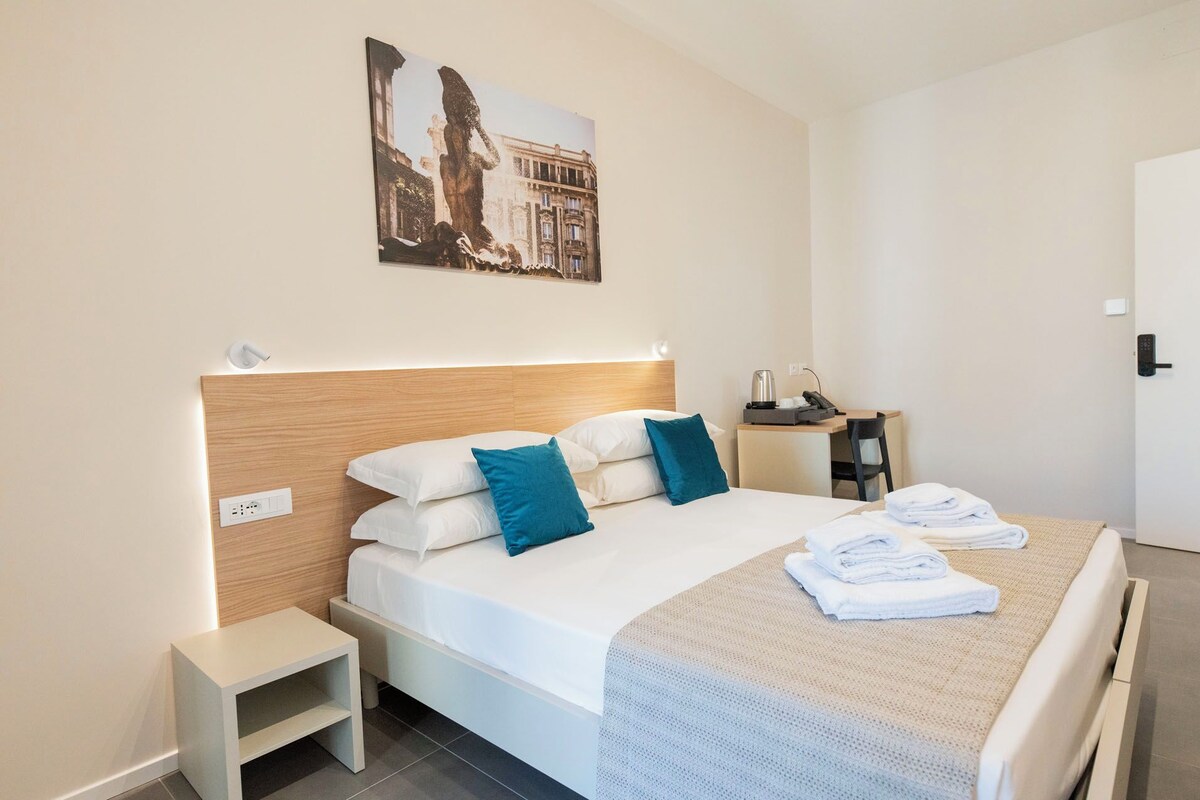 Una camera d'albergo con un letto, un comodino e una piccola scrivania con una sedia.