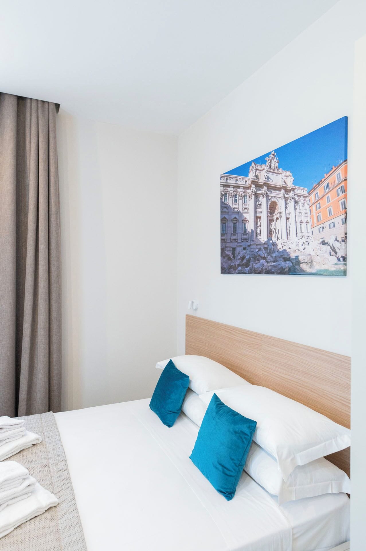 A hotel room with a bed, a headboard, a wall-mounted painting of a building, and a window with curtains.