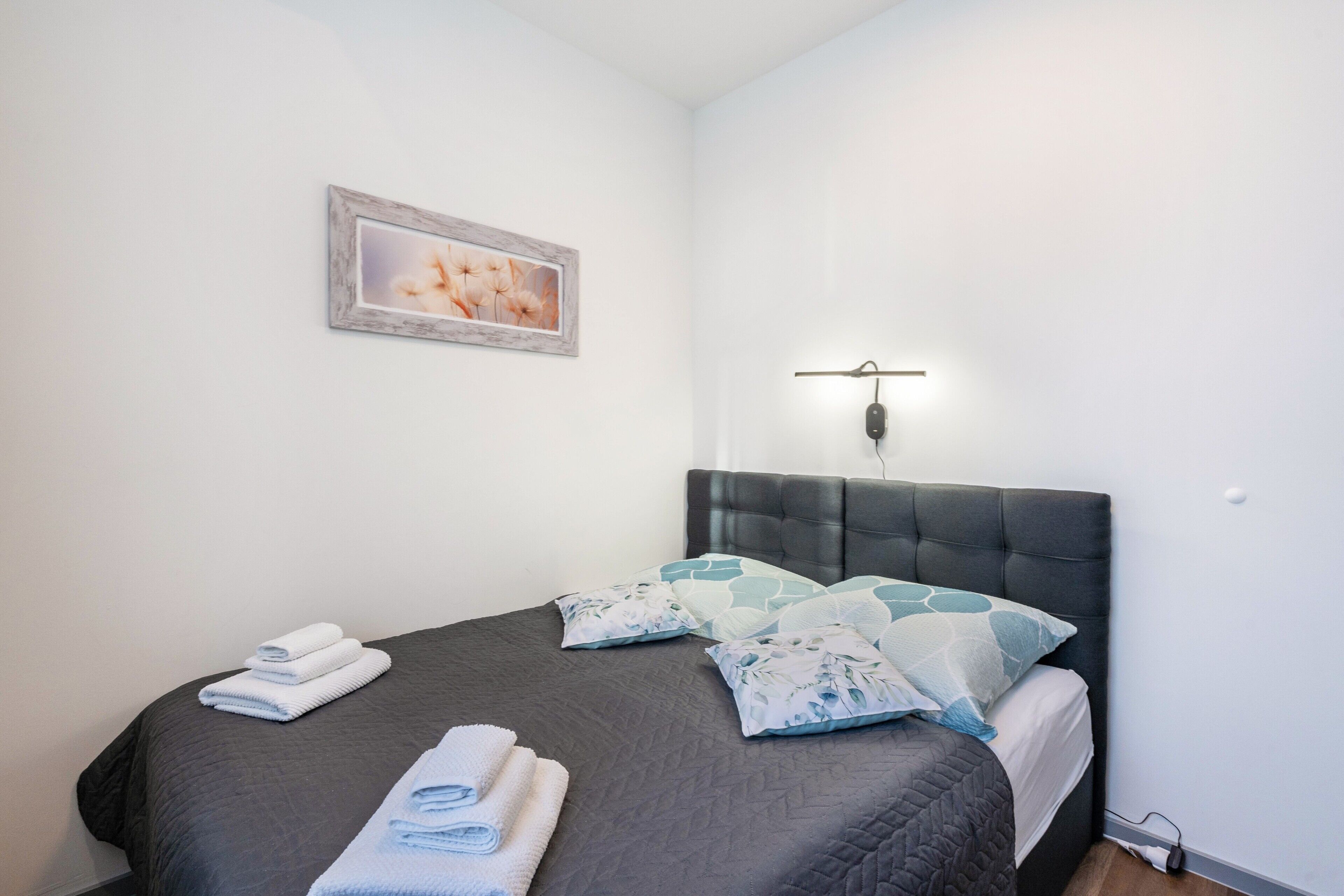 Un lit bien fait, avec des oreillers, une lampe fixée au mur et un tableau encadré.