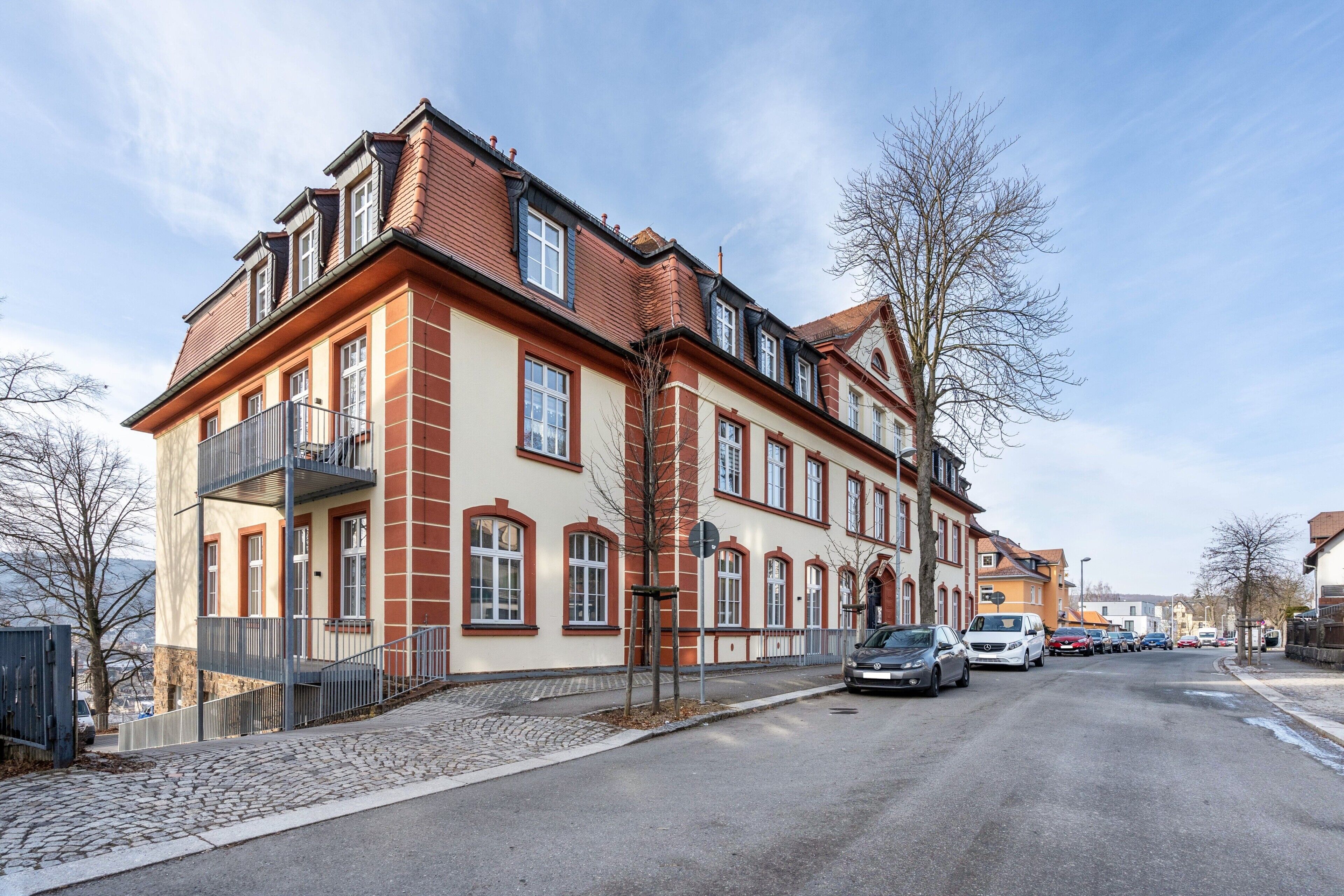 A multi-story building with a red and beige facade, a cobblestone street, and parked cars.