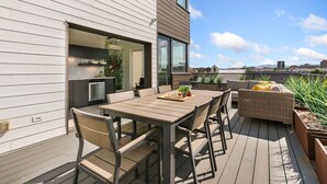 A modern outdoor dining area with a wooden table and chairs, a wicker sofa, and a kitchen visible through an open door. - The Overlook | Luxe Old Town Condo w/ King Suite (Fort Collins)