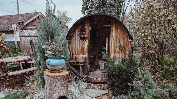 Une structure rustique en bois, surmontée d’un toit circulaire, entourée d’un jardin où poussent différentes espèces de plantes, et agrémentée d’une table en bois.