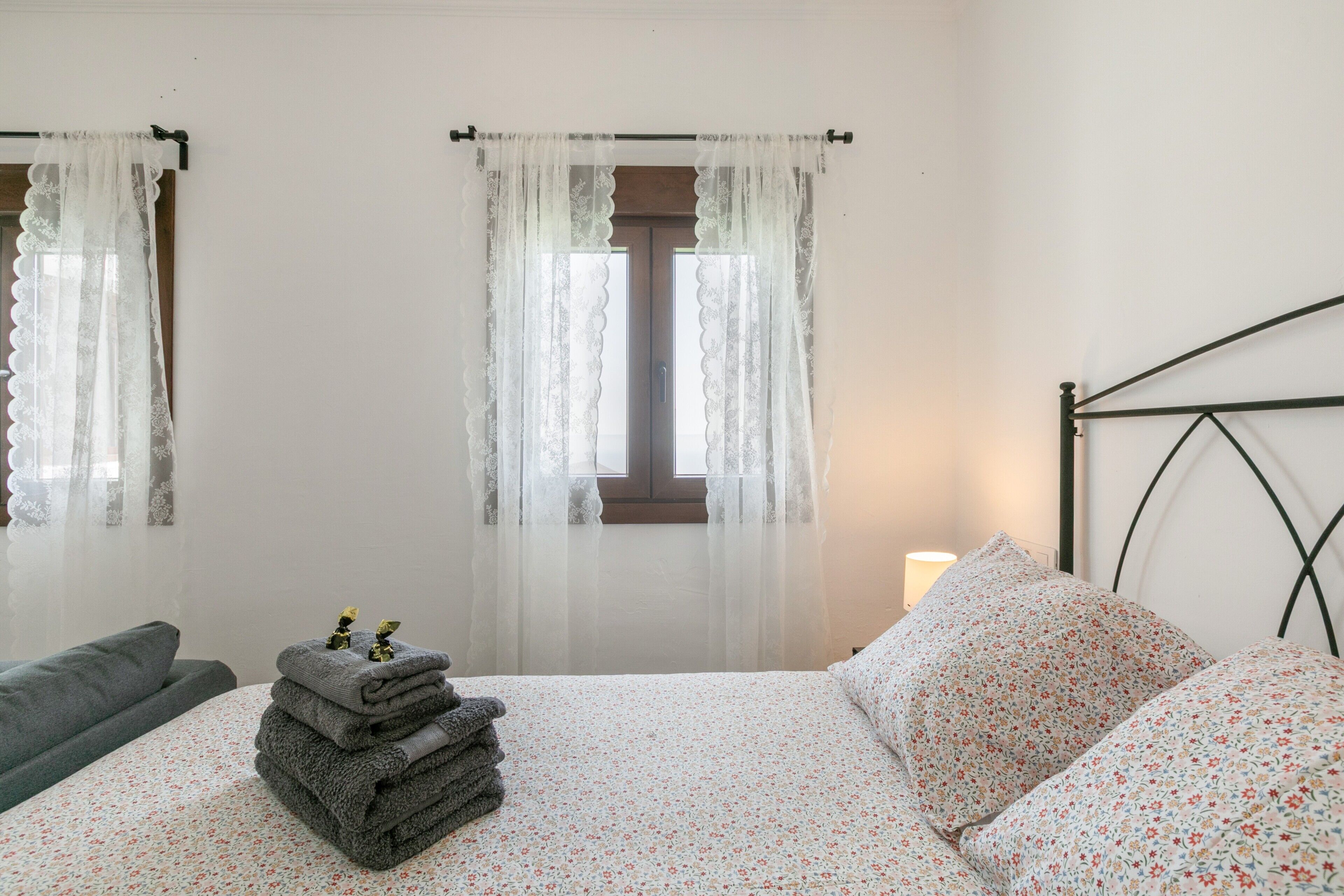 A bedroom with a bed, a stack of towels, a nightstand, and a window with sheer curtains.