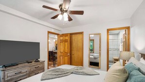 Une chambre à coucher avec un lit, un téléviseur posé sur une commode en bois, un ventilateur de plafond et une salle de bain visible à travers une porte ouverte.