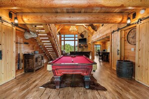 A wooden interior with a pool table, a chandelier, and a clock on the wall.