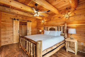Une chambre dans une cabane en bois, avec un lit, un ventilateur de plafond et une table de chevet avec une lampe.