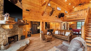 A log cabin interior with a stone fireplace, leather furniture, a pool table, and a mounted deer head.