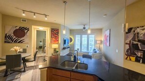 A modern kitchen with a black countertop, stainless steel sink, and a dining area with a table and chairs. There are abstract paintings on the walls and a large window with blinds.