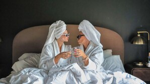 Dos personas con batas sentadas en una cama con un cabecero grande, sosteniendo copas.