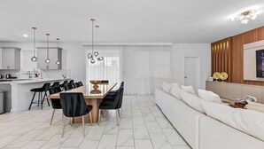 A modern living room with a white sofa, a dining table with black chairs, and a kitchen area with white cabinets and pendant lights.