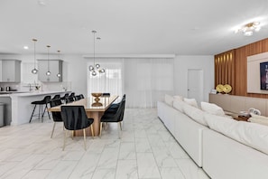 Una sala moderna con un sofá blanco, una mesa de comedor con sillas negras y una zona de cocina con armarios blancos y luces colgantes.