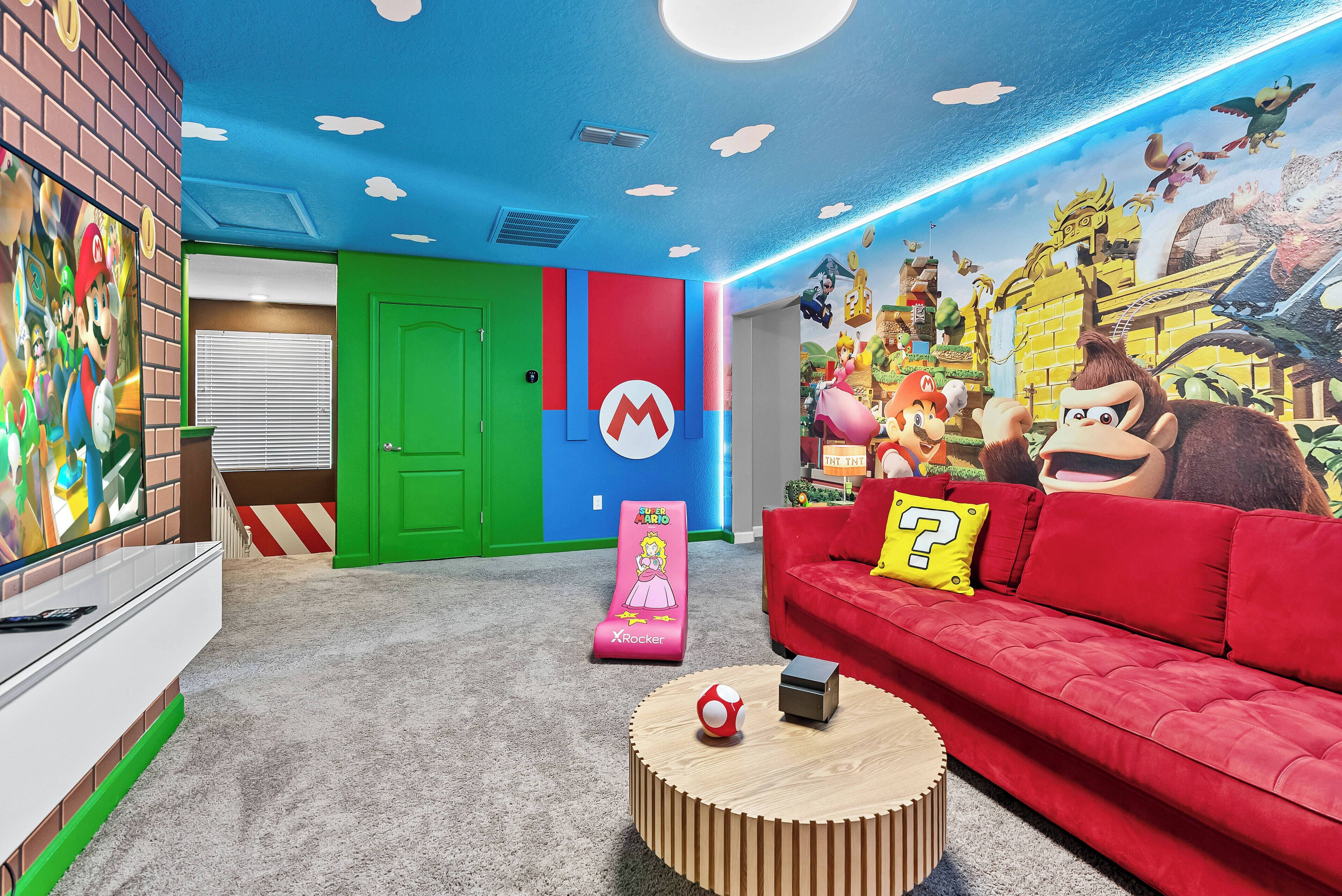 Una habitación vibrante con un mural temático de Nintendo, un sofá rojo con un cojín amarillo y una Nintendo DS sobre una mesa redonda de madera.