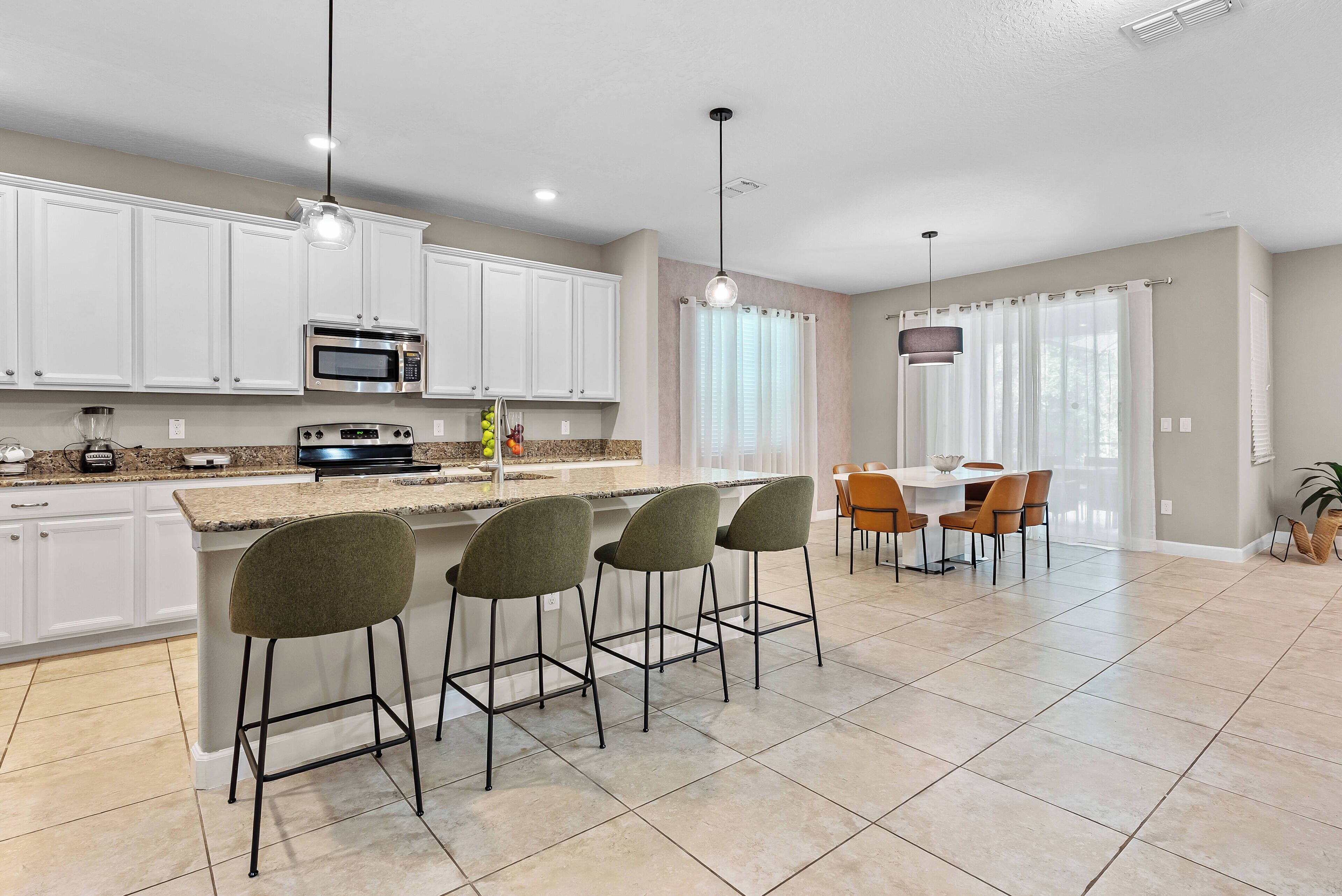 A modern kitchen with white cabinets, a central island with bar stools, a dining area with a table and chairs, and a large window with sheer curtains.
