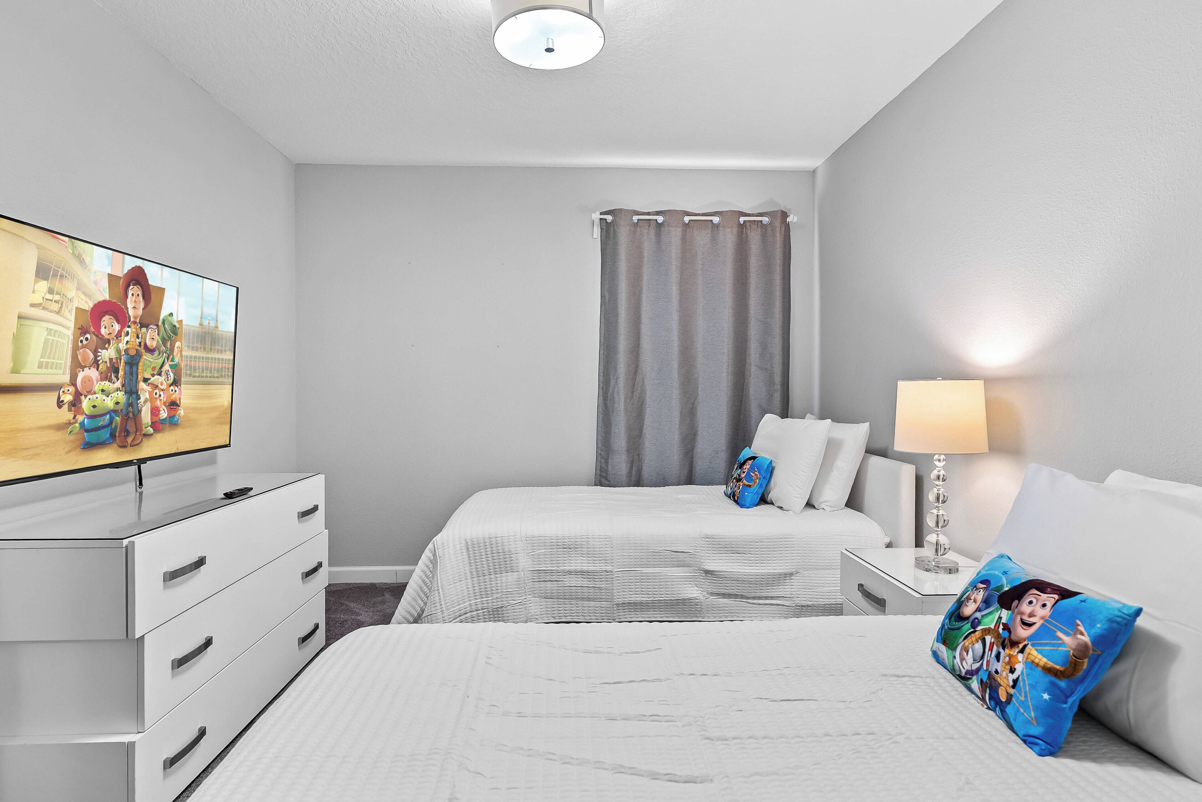 Una habitación de hotel con televisión de pantalla plana, una cama con ropa blanca, una mesita de noche con lámpara y una ventana con cortina gris.