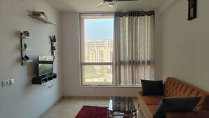 A living room with a sofa, a TV mounted on the wall, a ceiling fan, and a large window with curtains. - Spacious 1BHK Hiranandani Estate (Thane)