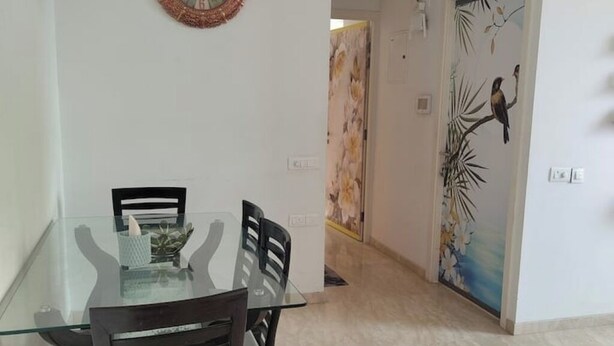 A dining area with a glass table and wooden chairs, a wall clock, and a door with a bird mural. - Spacious 1BHK Hiranandani Estate (Thane)